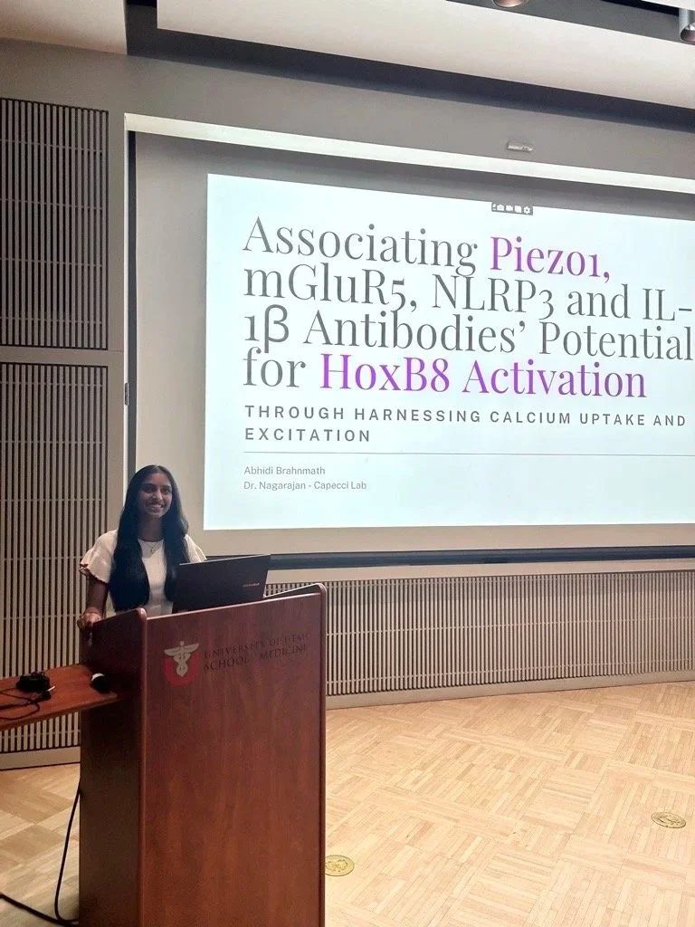 A woman stands at a podium in front of a projection screen displaying a scientific presentation titled 'Associating Piezo1, mGluR5, NLRP3 and IL-1β Antibodies' Potential for HoxB8 Activation'. The podium has a symbol and text indicating it is from the University of Illinois College of Medicine.