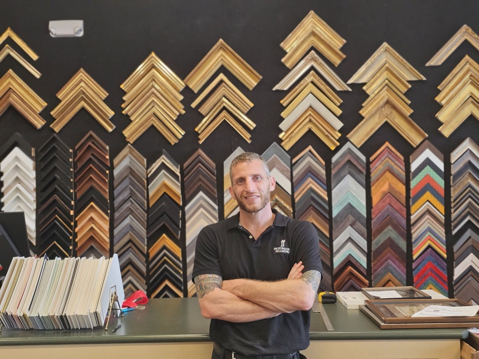 Adam Fins, owner of Friedman's Framing in Savannah, Georgia, standing with arms crossed at the shop counter in front of a wall of frame corner samples