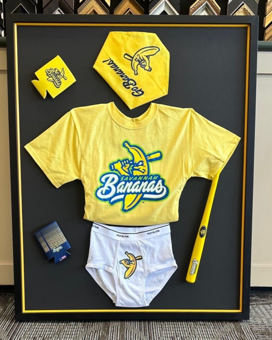 A black shadow box framing a yellow Savannah Bananas T-shirt, cap, bat, koozies and branded underwear