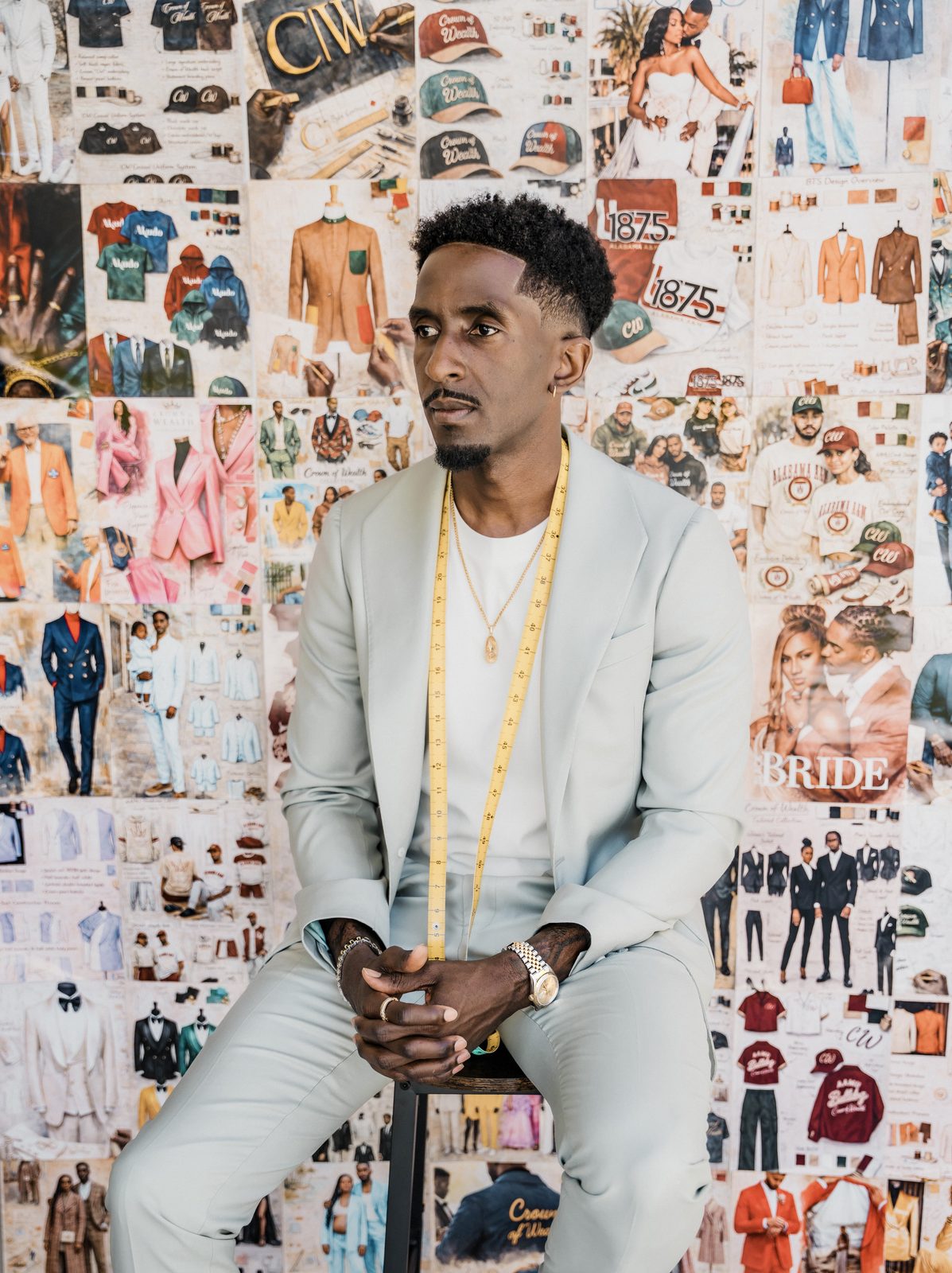 Adeola Odutola, founder and creative director of Crown of Wealth, seated in a pale suit with a tape measure around his neck in front of a wall of his designs