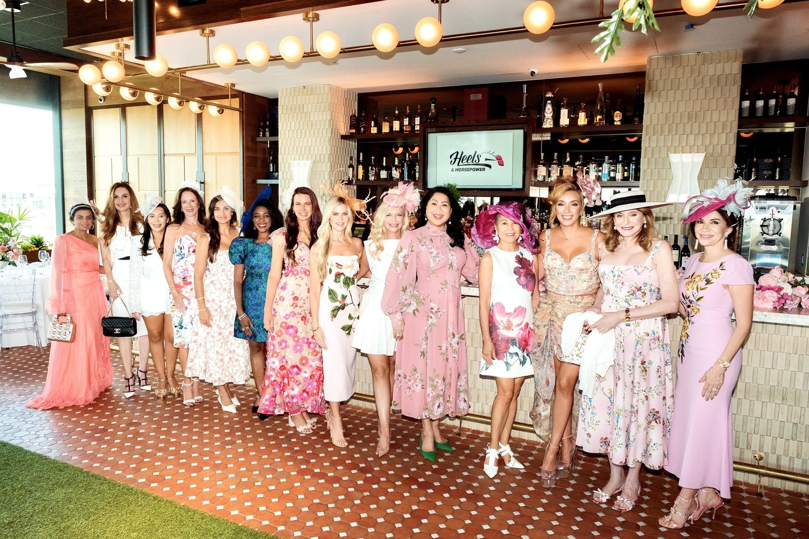 Fifteen Heels & Horsepower members in floral dresses and statement hats lined up in front of a bar with the club logo on screen