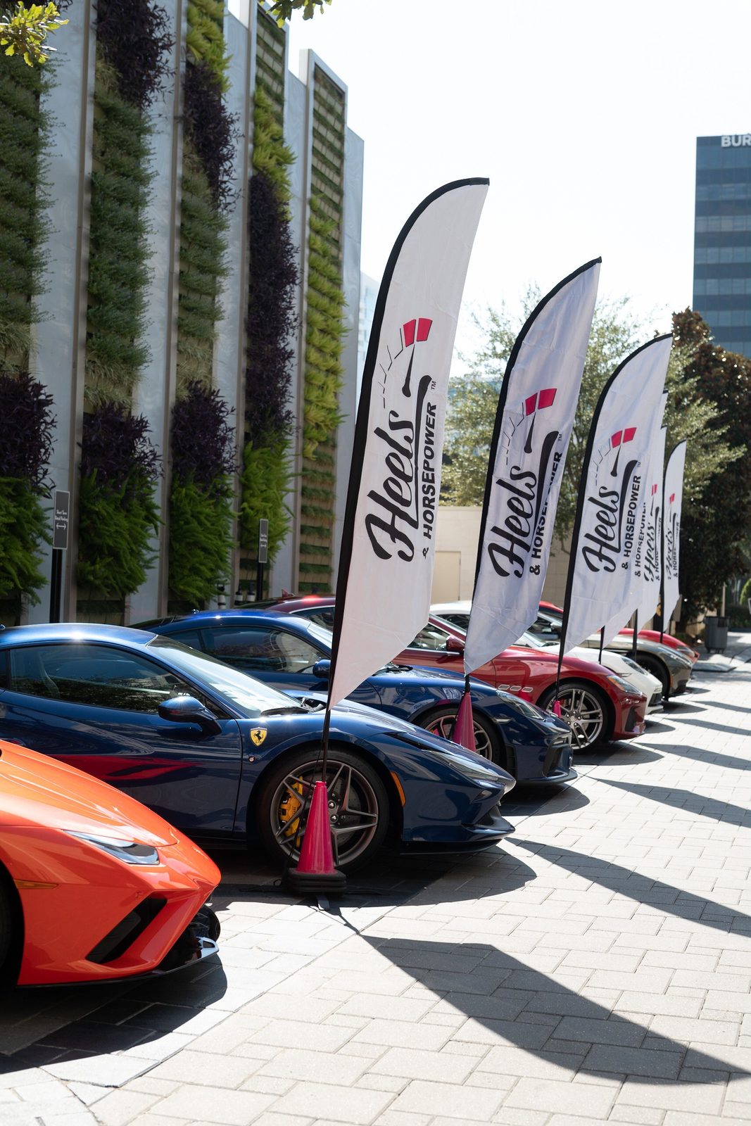 A row of exotic cars, including a blue Ferrari and an orange supercar, parked beneath Heels & Horsepower feather flags beside a green wall
