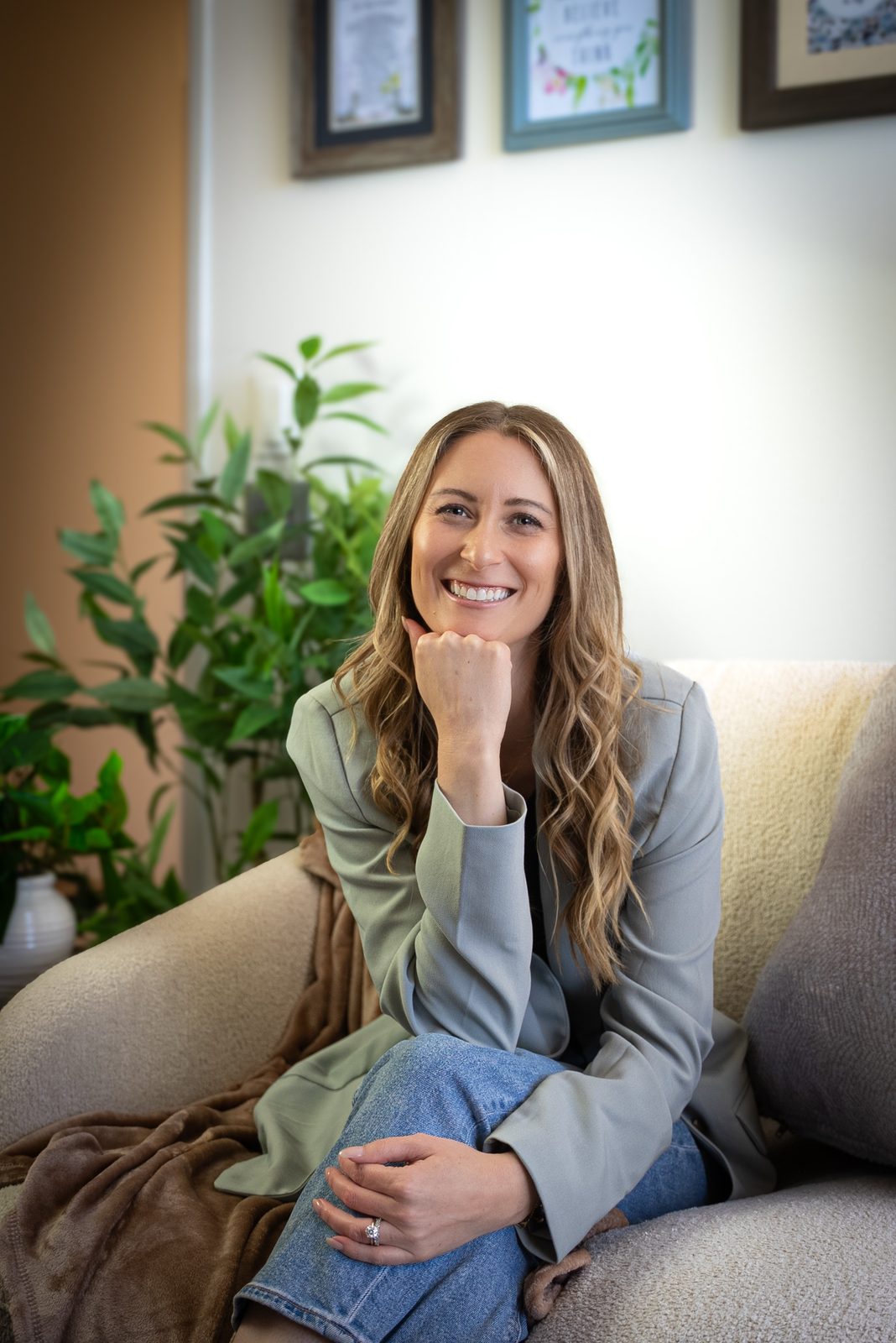 Alexis Flores smiling on a cream sofa in her therapy office, chin resting on her hand, beside a leafy green plant