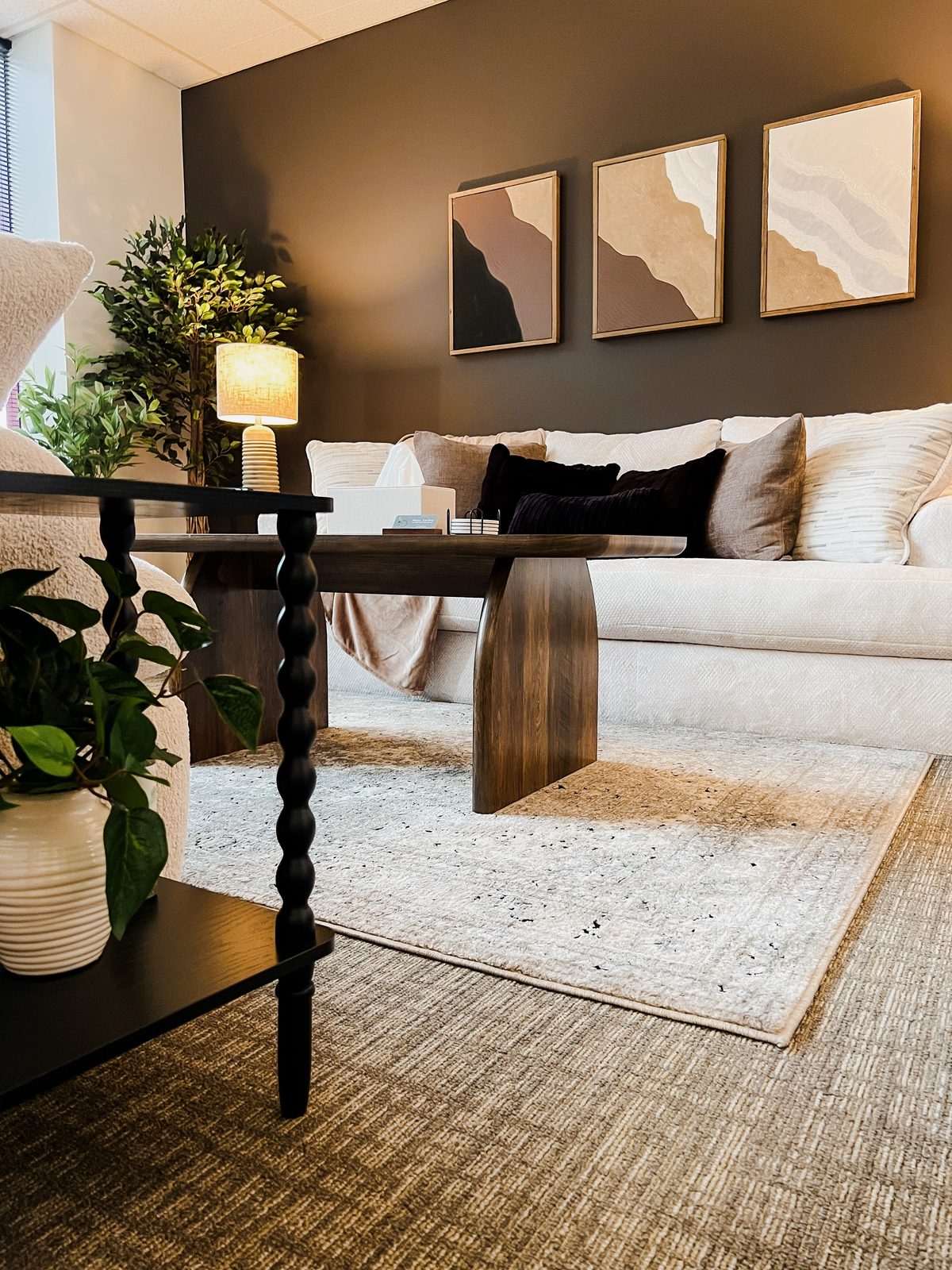 A Therapy Worthwhile therapy room with a white sofa, a wooden coffee table, a lamp and three abstract paintings on a dark wall