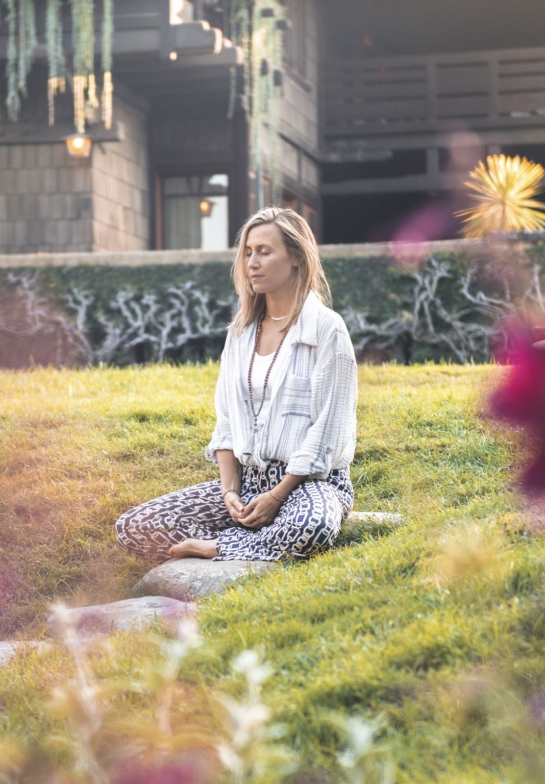 Anna Christine Bowden meditating cross-legged on a grassy slope in front of a shingled craftsman house