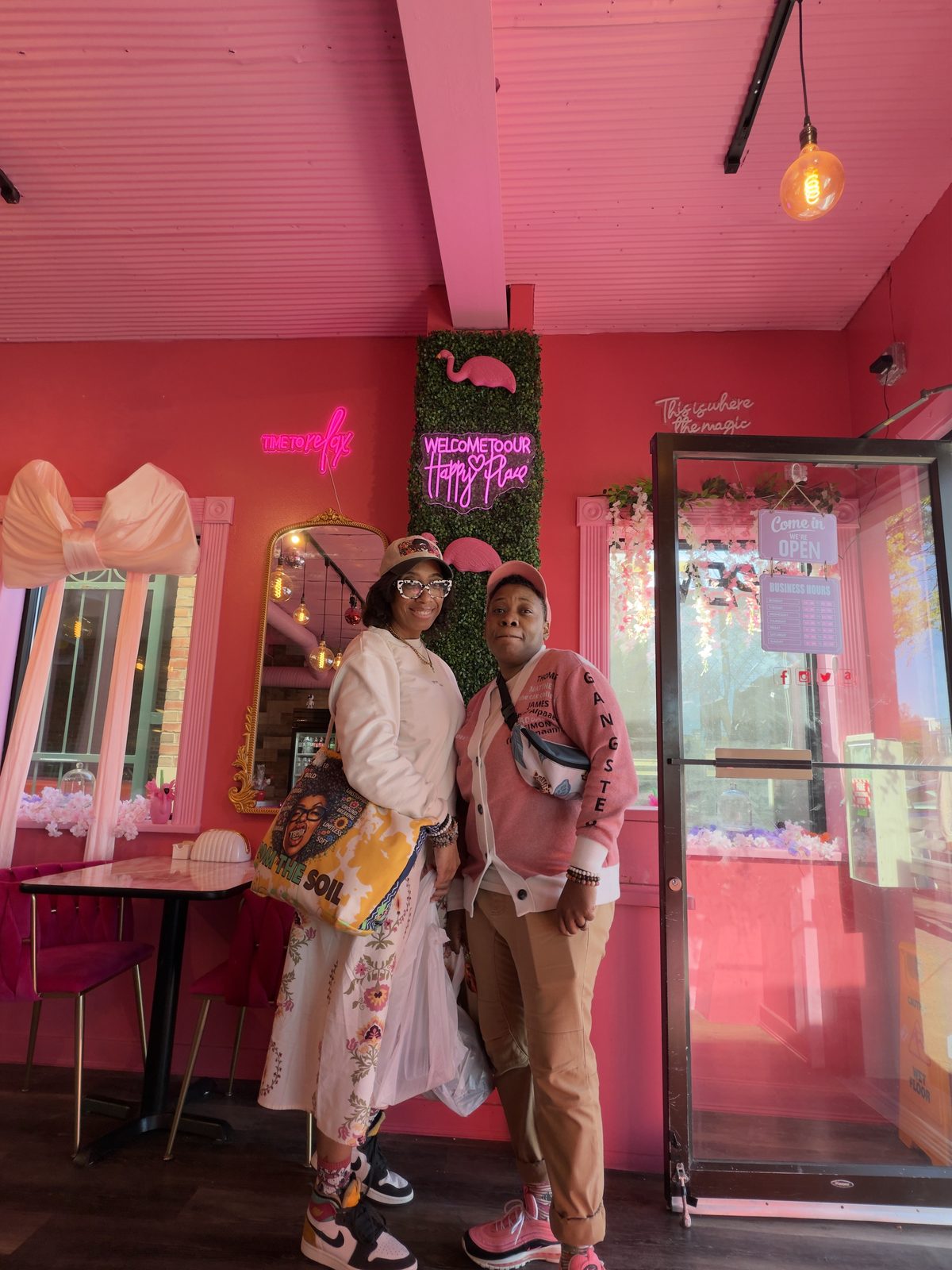 Arlesia and Letecia Whitaker, co-founders of TBOM Apparel, together inside a pink cafe beneath a neon Welcome to Our Happy Place sign