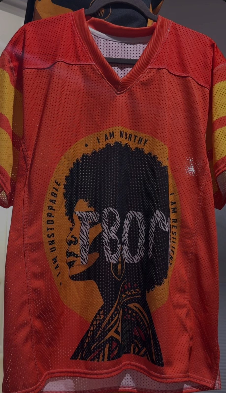 Red TBOM Apparel mesh jersey with the brand’s afro silhouette design ringed by the words I Am Worthy, I Am Unstoppable and I Am Resilient