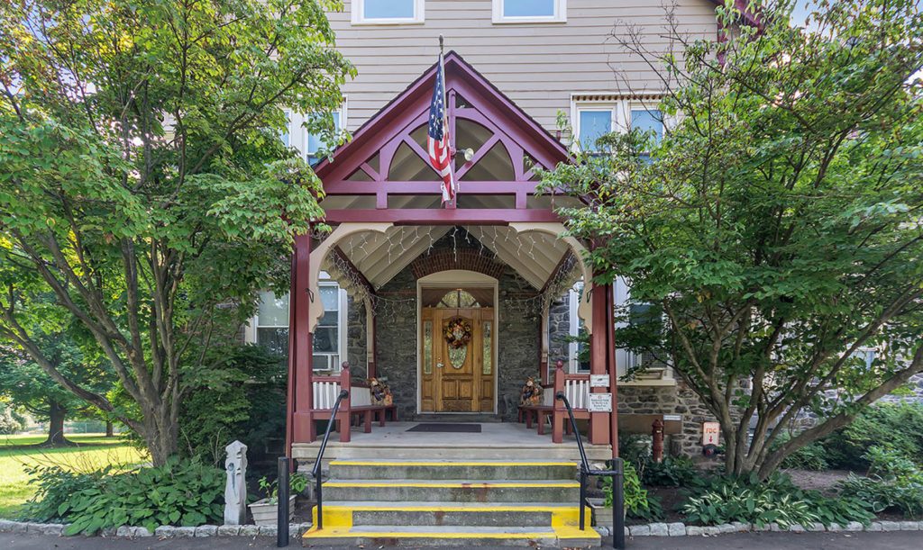 The front porch of Roxborough Home for Women, a stone and clapboard house with a maroon gabled entrance and an American flag