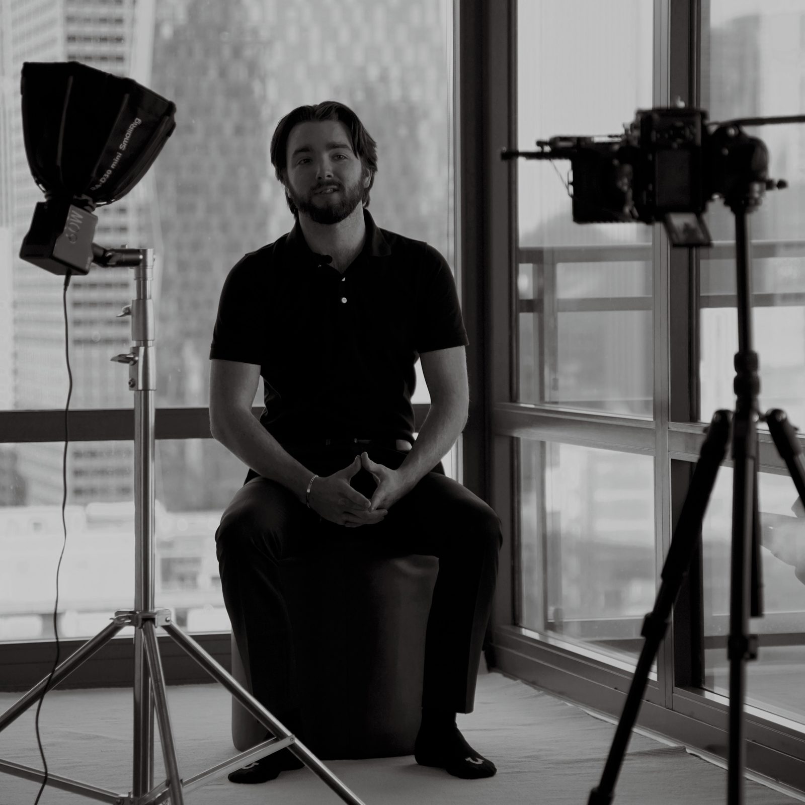 Austin Coburn seated on a stool between a softbox light and a cinema camera on a tripod, with the downtown Chicago skyline behind the windows