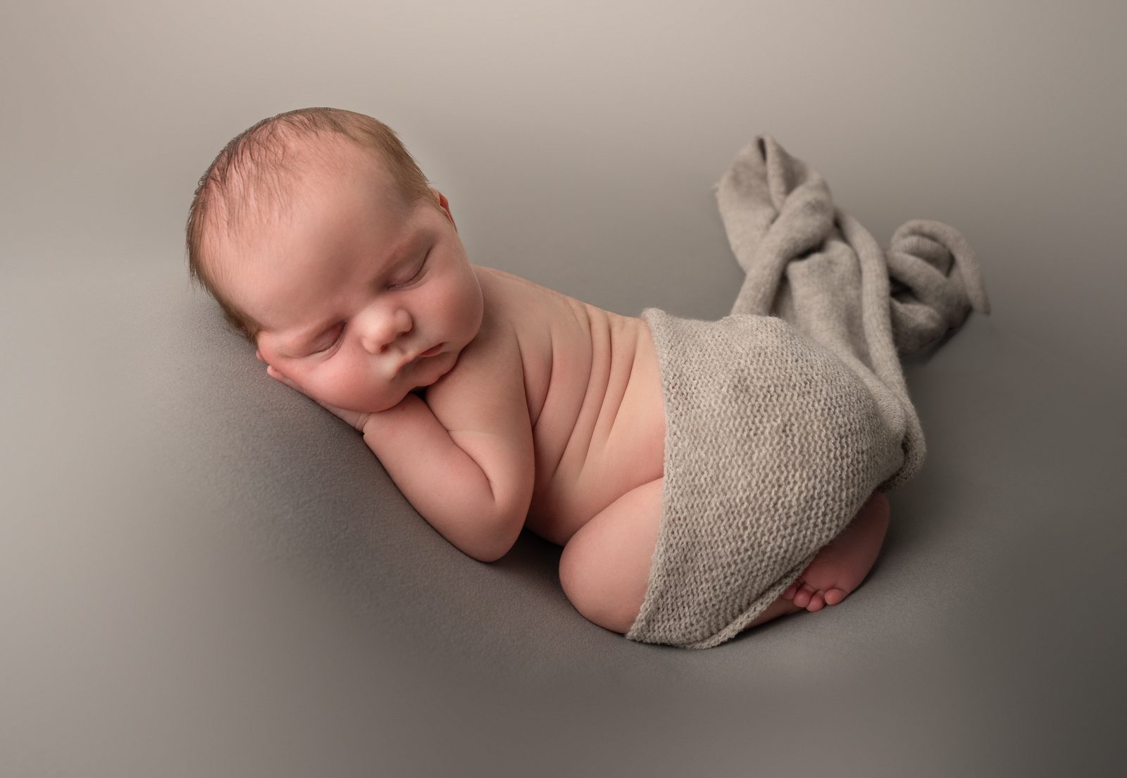 A newborn asleep on its side under a grey knit wrap, photographed by Mess Of A Dreamer Photography
