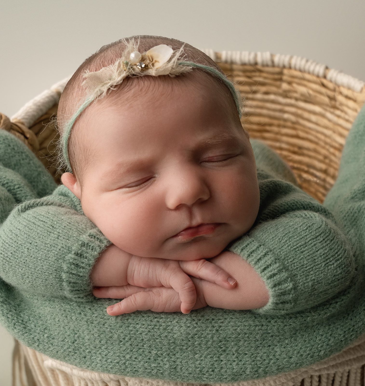 A sleeping newborn in a green knit romper and floral headband, resting its chin on its hands in a woven basket