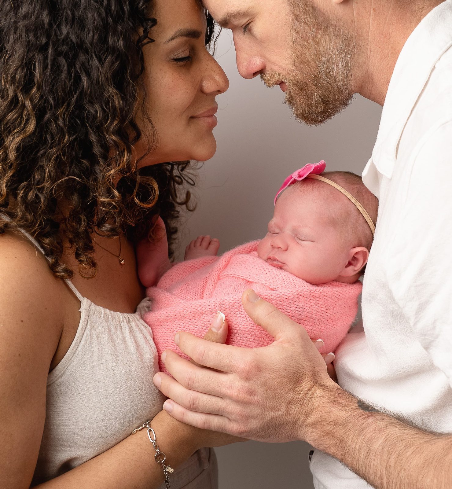 New parents leaning in forehead to forehead over their sleeping baby girl, swaddled in pink
