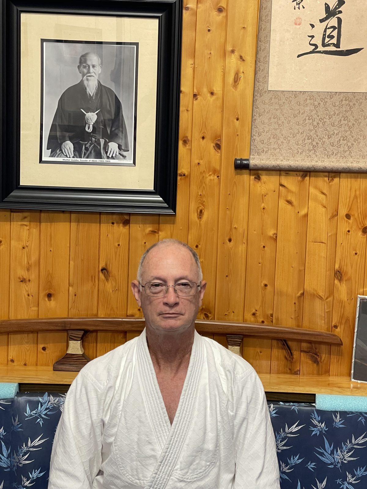 Bill Hague in a white aikido gi, seated in front of a wood-paneled wall beneath a framed portrait of the founder of aikido