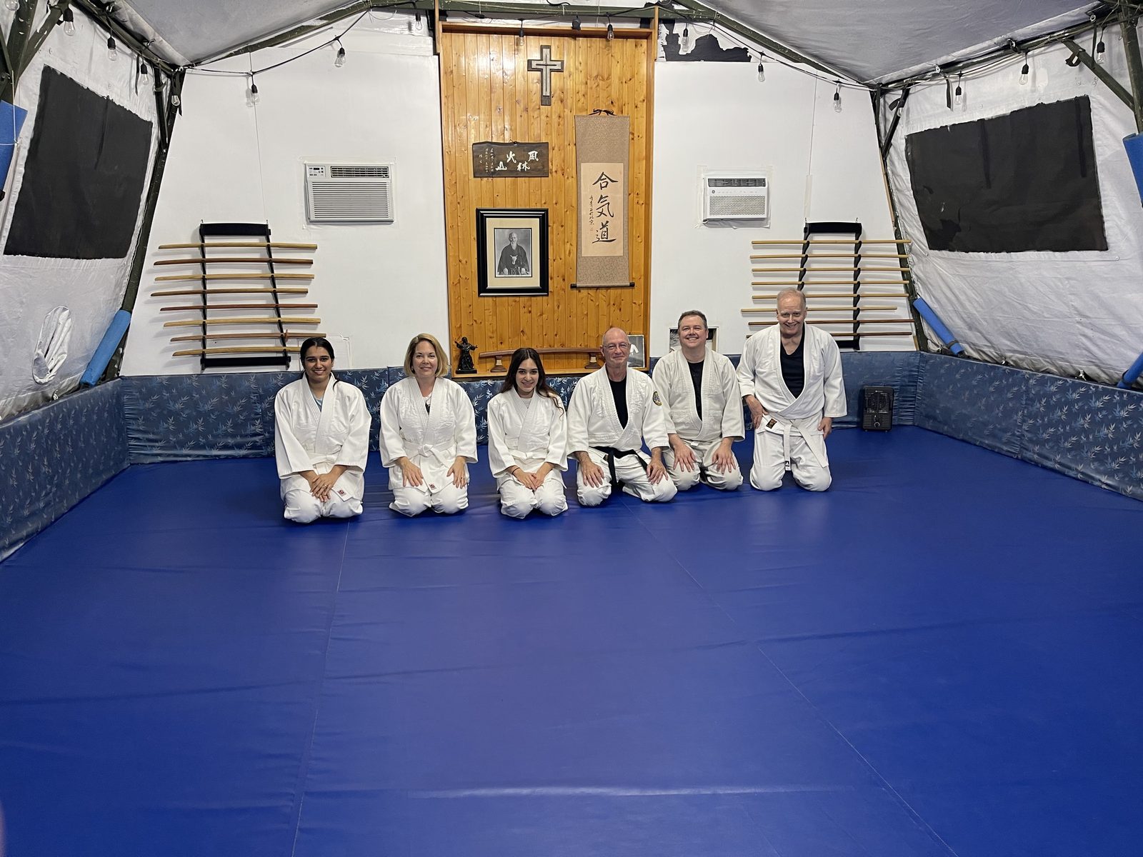 Six aikido students and instructors in white gis kneeling in a row on the blue mat at Mushin Dojo