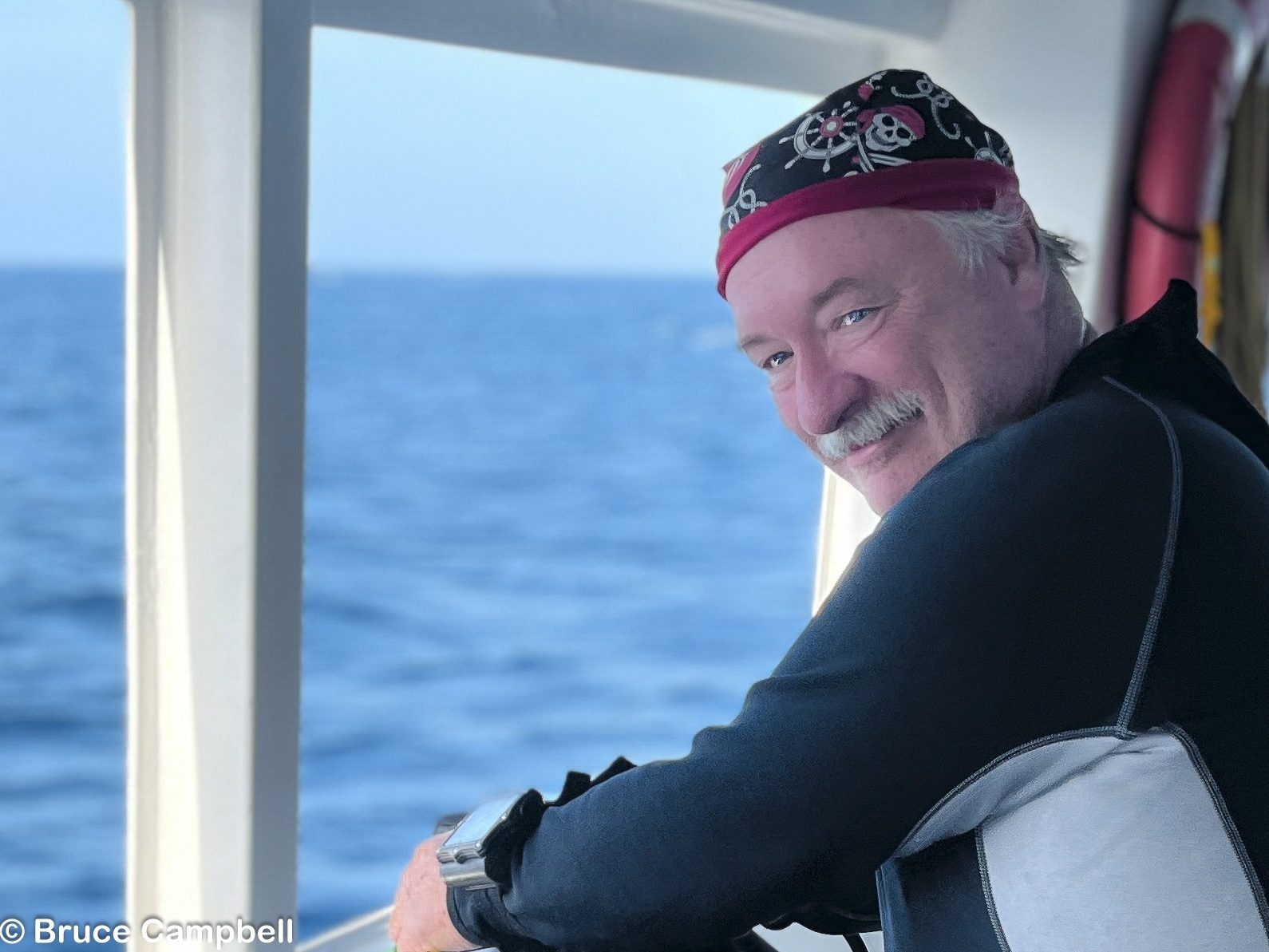 Bruce Campbell, founder of Bruce Campbell Photography, smiling at the rail of a dive boat in a wetsuit and a skull-print bandana