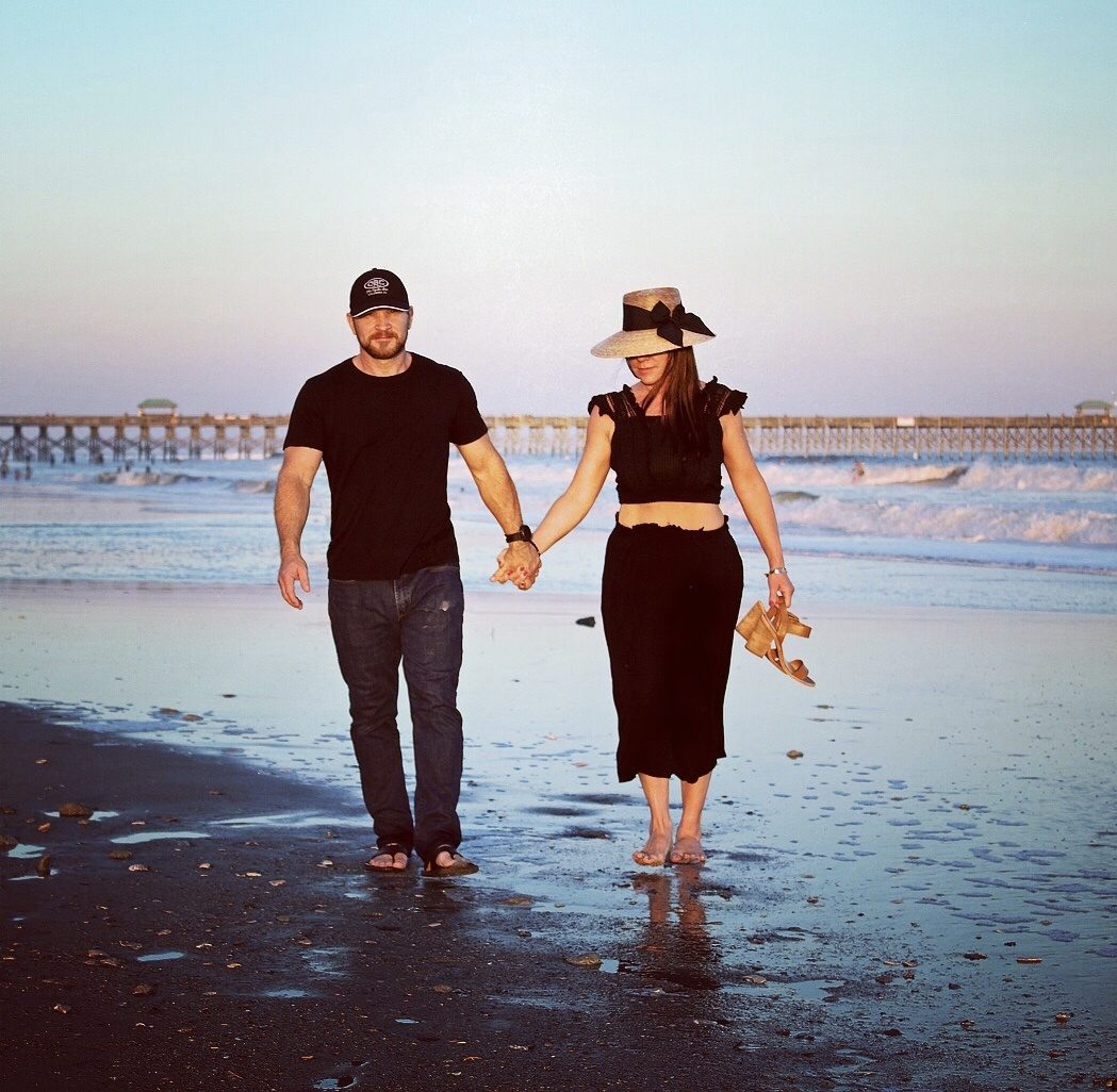 Carmen Holley in a straw hat walking the beach at sunset with her husband, holding hands near a pier