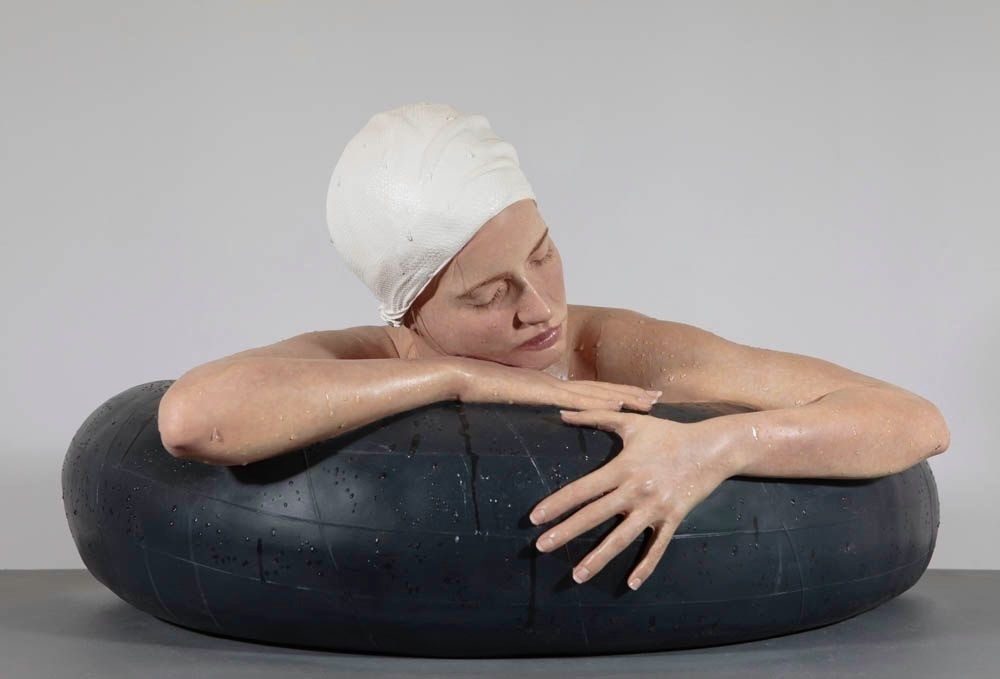 A Carole Feuerman superrealist sculpture of a swimmer in a white cap resting her arms and head on a black inner tube