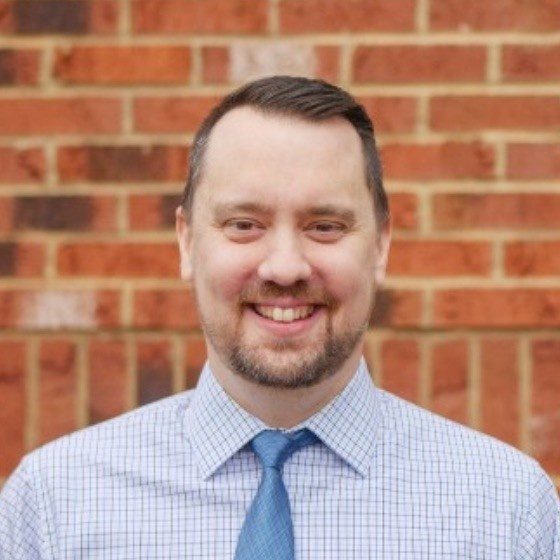 Casey Carmical, founder of 1-FIND SERVICES, smiling in a checked shirt and blue tie in front of a red brick wall