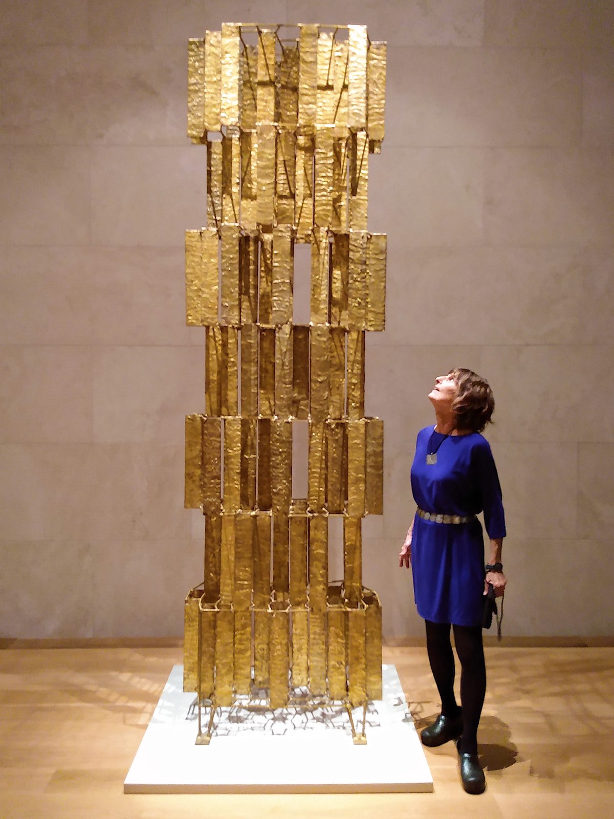 Celia Bertoia in a blue dress looking up at a tall golden Harry Bertoia sculpture in a museum gallery