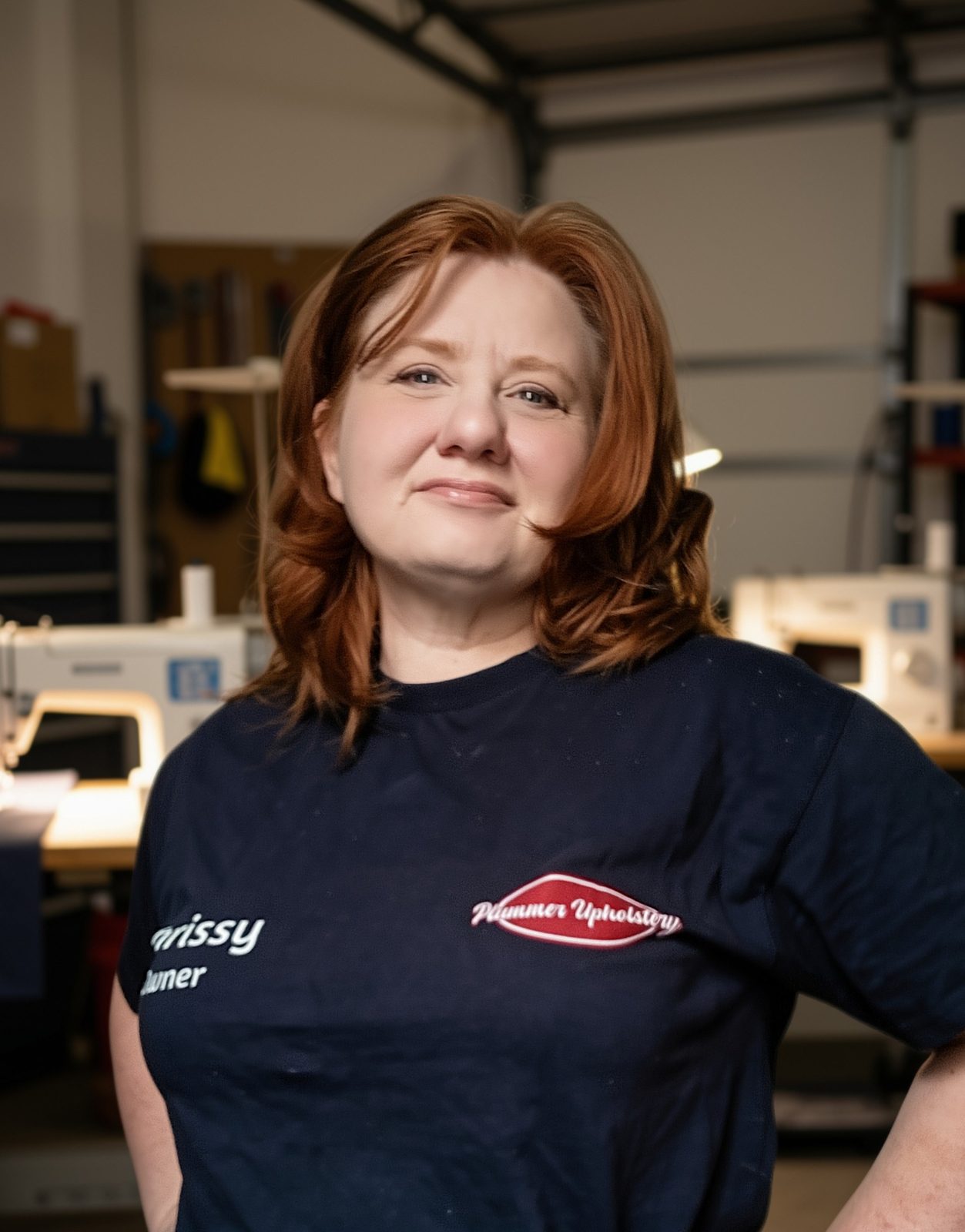 Christena Plummer, founder of Plummer Upholstery, in her workshop in Fallston, Pennsylvania, wearing a navy shop shirt with the Plummer Upholstery logo, sewing machines behind her