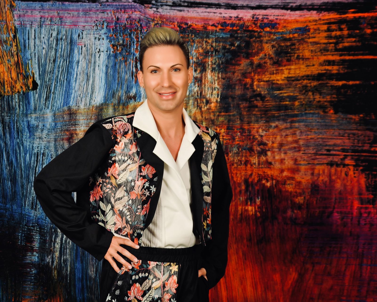 Corey Locke, founder of Aristotle Cosmetics, in a white shirt and floral-embroidered black jacket in front of a painted backdrop