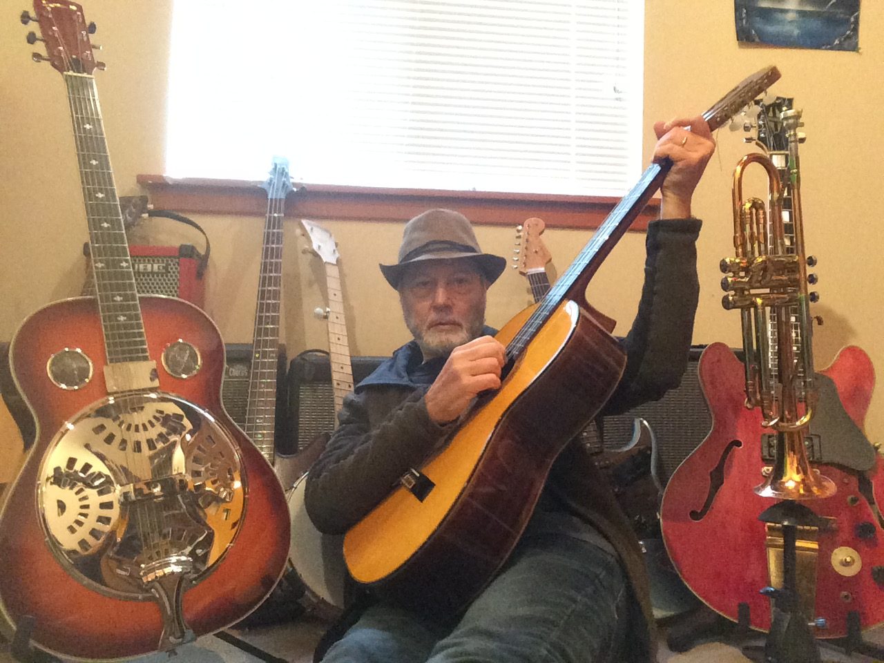 Dan Zalles in a hat at home with an acoustic guitar, surrounded by a resonator guitar, a bass, an electric guitar and a trumpet