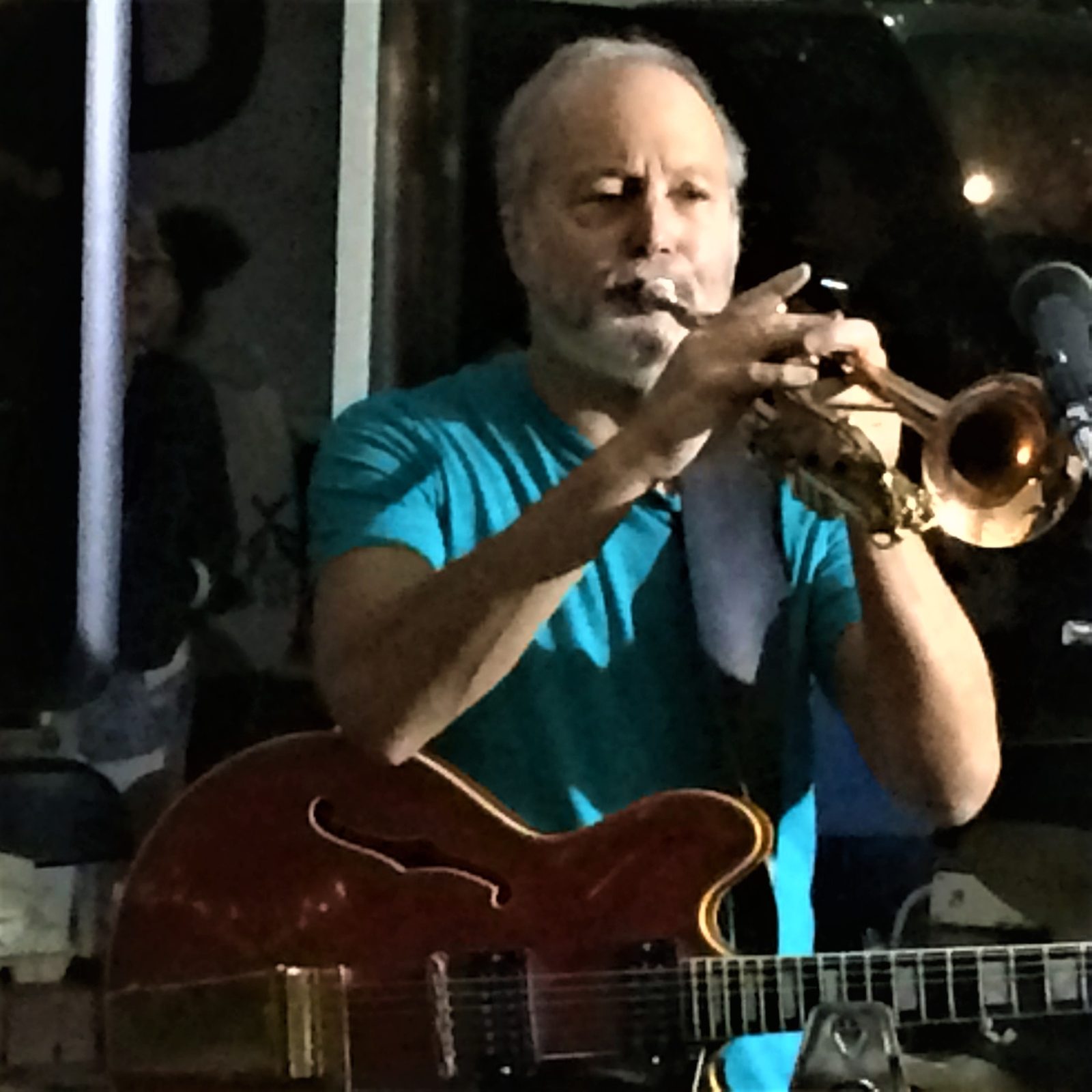 Dan Zalles in a teal shirt playing trumpet on stage with a red electric guitar slung in front of him