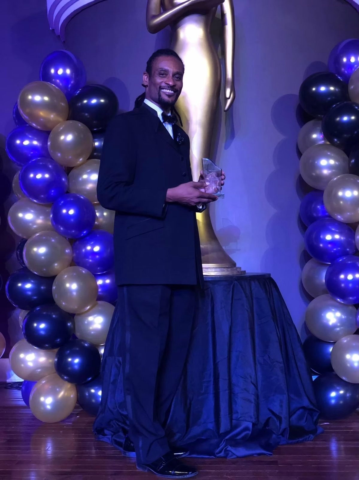Dearick Milton in a tuxedo holding his GCMA award beside gold and purple balloons