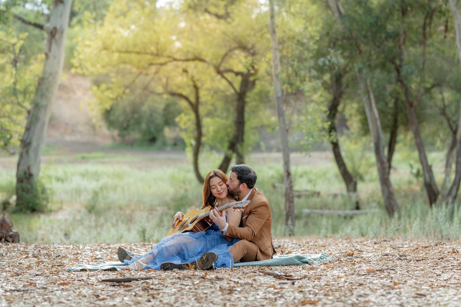 A couple sharing a guitar on a picnic blanket among the trees, photographed by Divya Stumpff