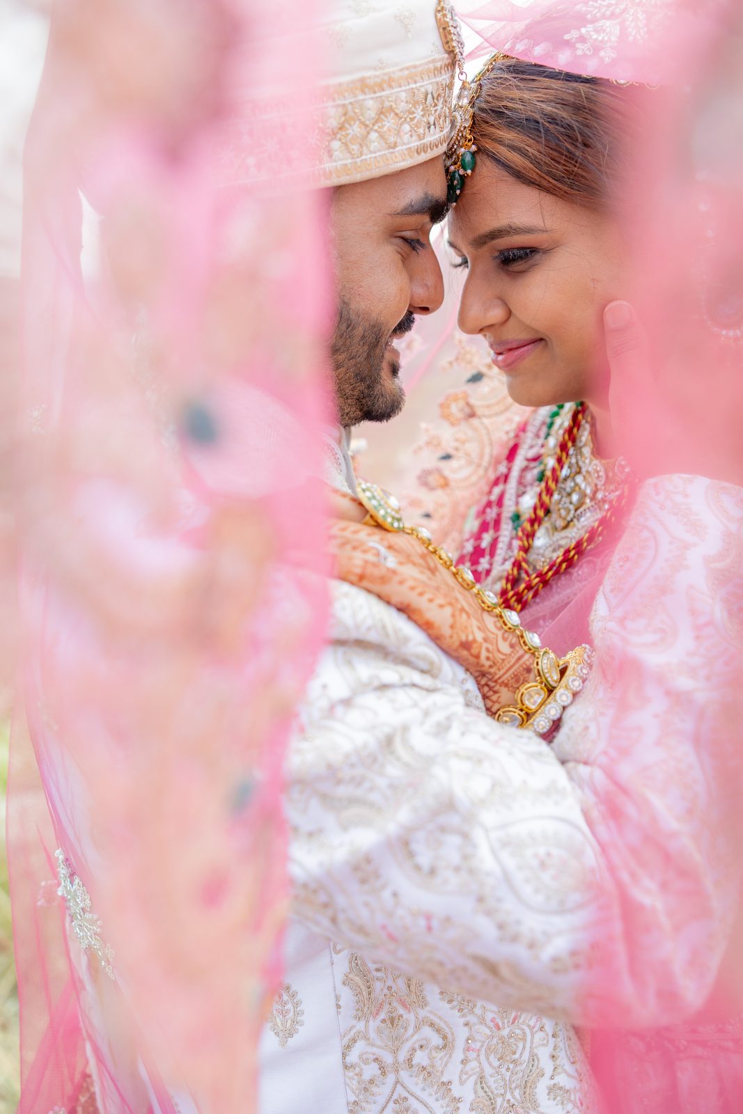 A couple in traditional Indian wedding attire framed by a pink veil, photographed by Divya Stumpff