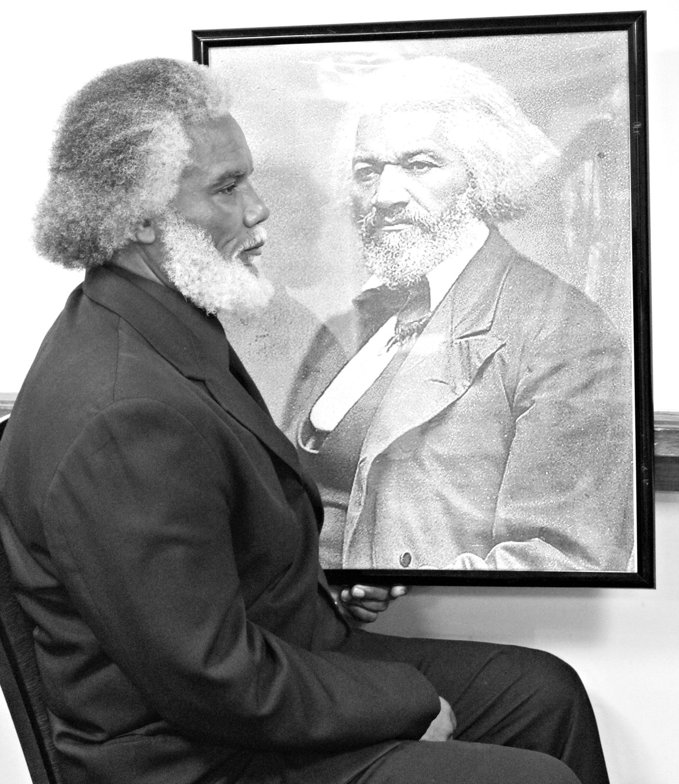 Donald L. Dowridge, Jr. in character as Frederick Douglass, seated beside a framed portrait of Douglass
