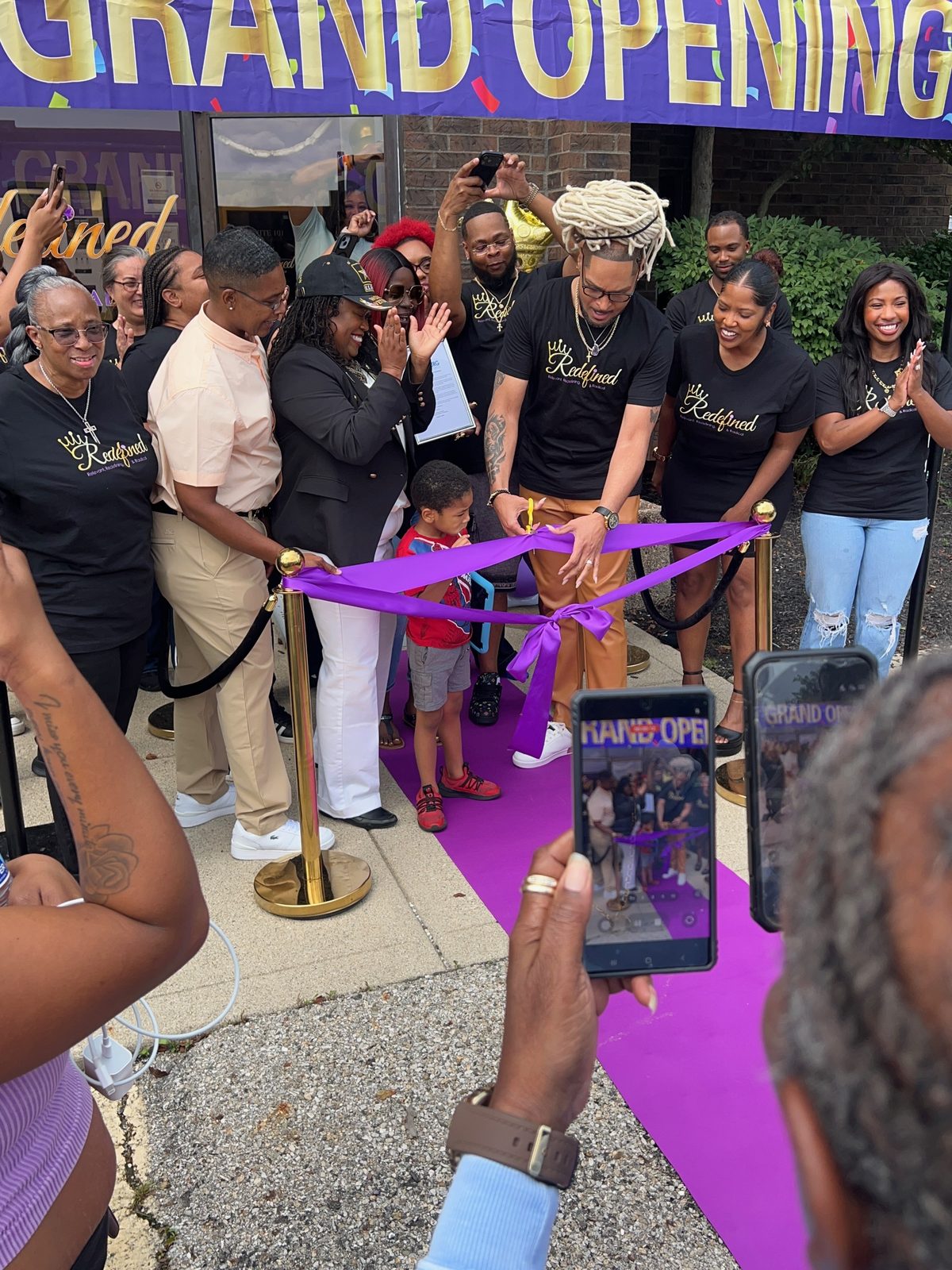 A crowd cheering a purple ribbon cutting under a Grand Opening banner at Redefined