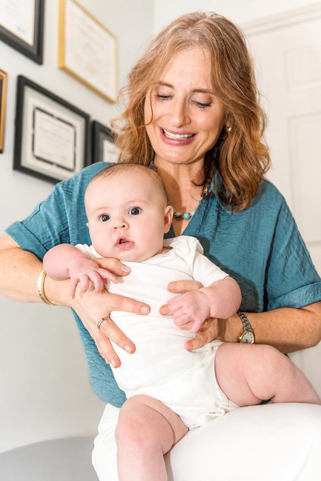 Elizabeth Geiger Harvey holding a baby in her office, framed diplomas on the wall behind her