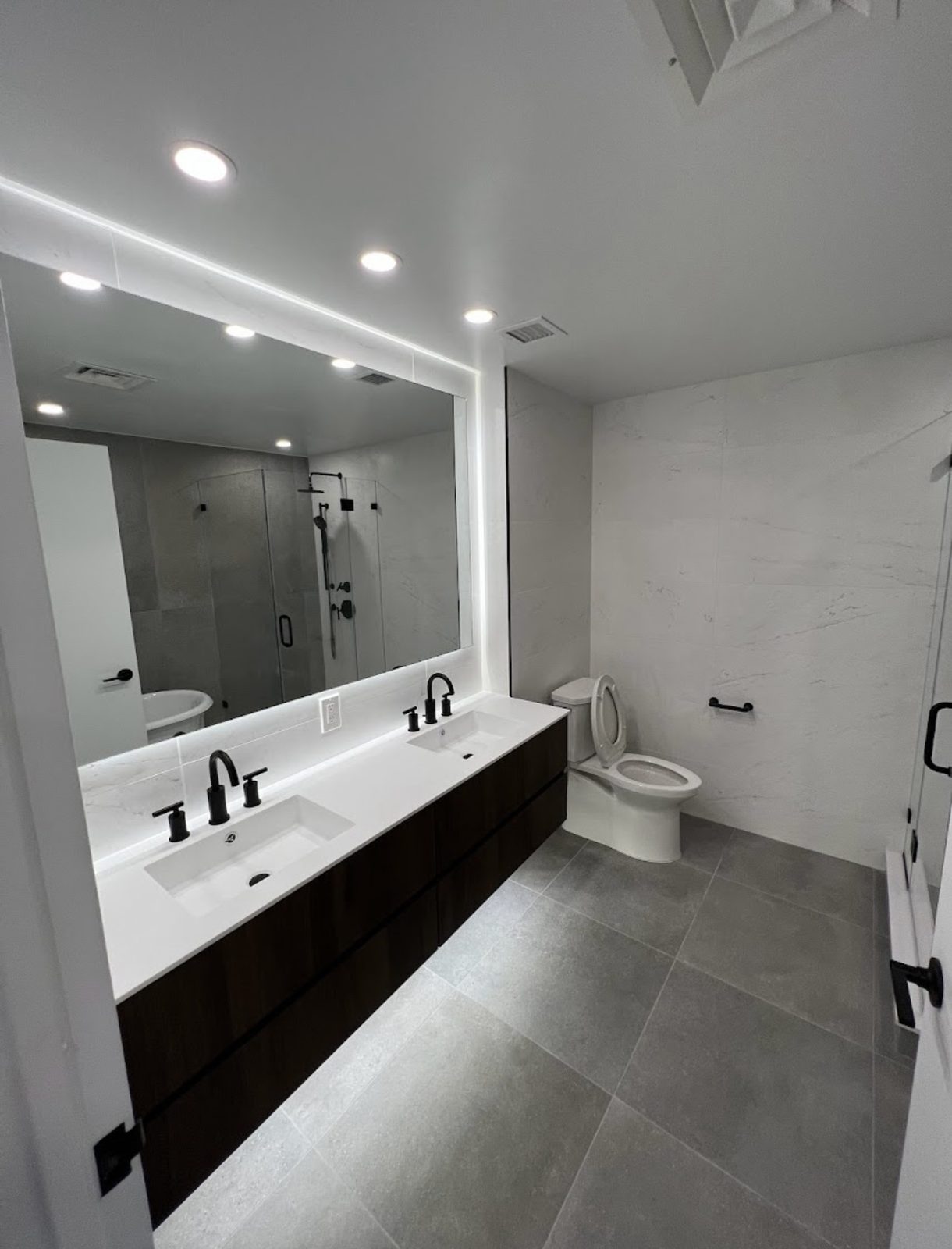 A renovated bathroom by Mega Group with a backlit mirror, double vanity, matte black fixtures and large grey floor tiles
