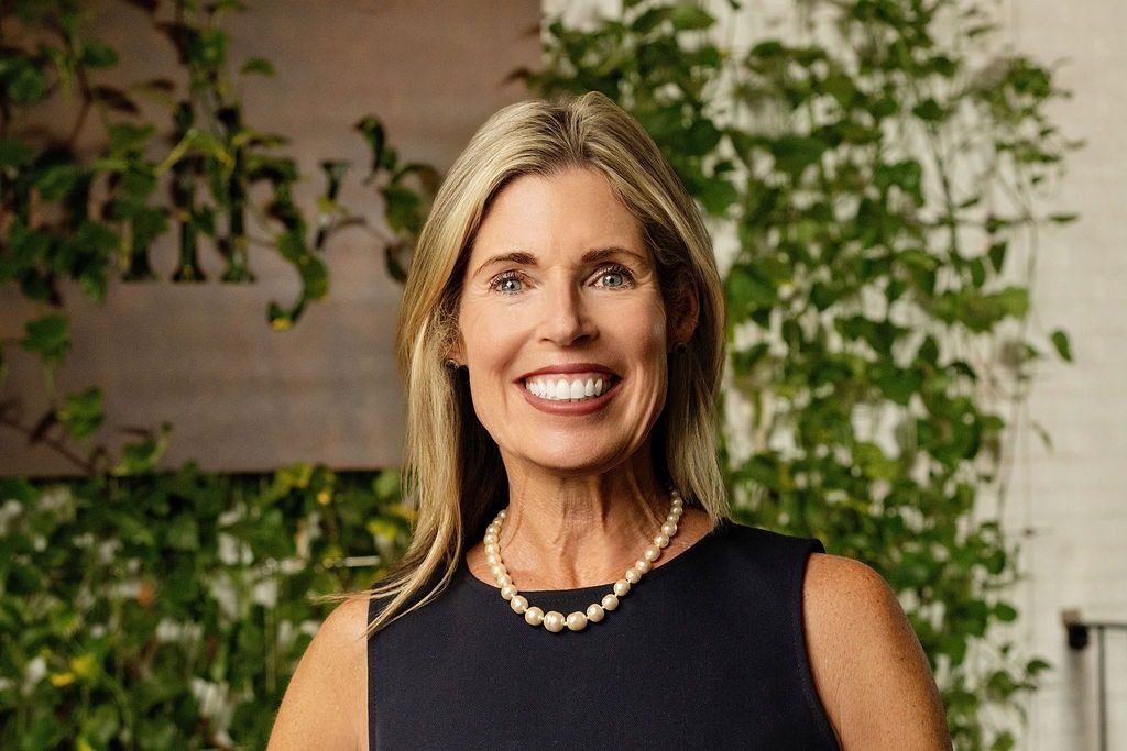 Emily Fournier, founder of Emily’s Table, smiling in a navy dress and pearl necklace in front of a green ivy wall