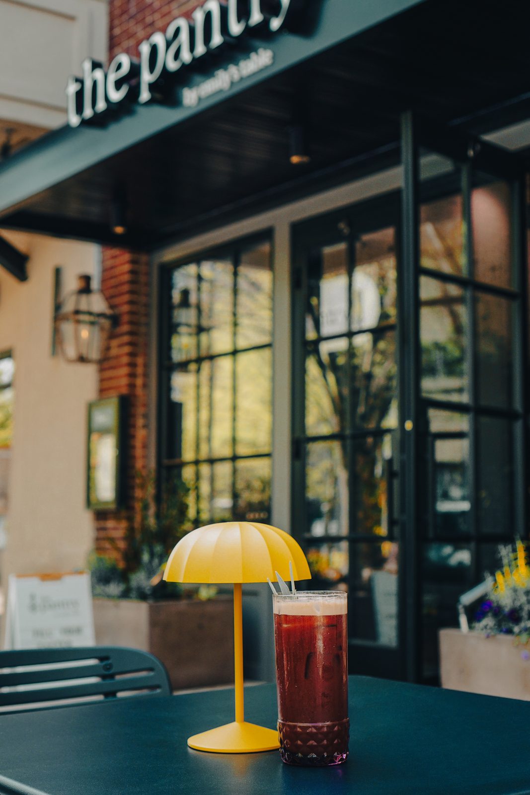 A tall iced red drink beside a yellow table lamp on a sidewalk table in front of the Pantry