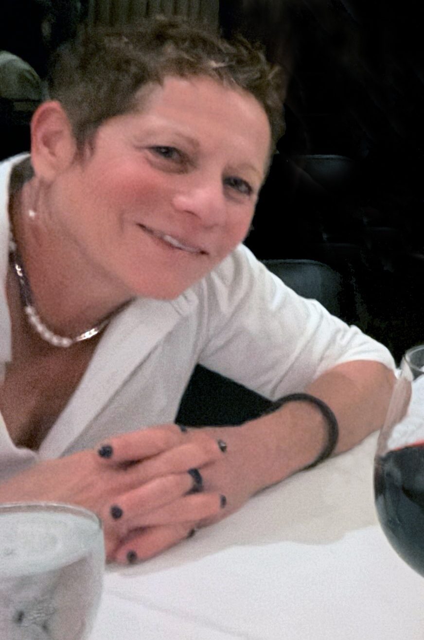 Emily Goldstein smiling at a restaurant table in a white shirt, a glass of red wine beside her