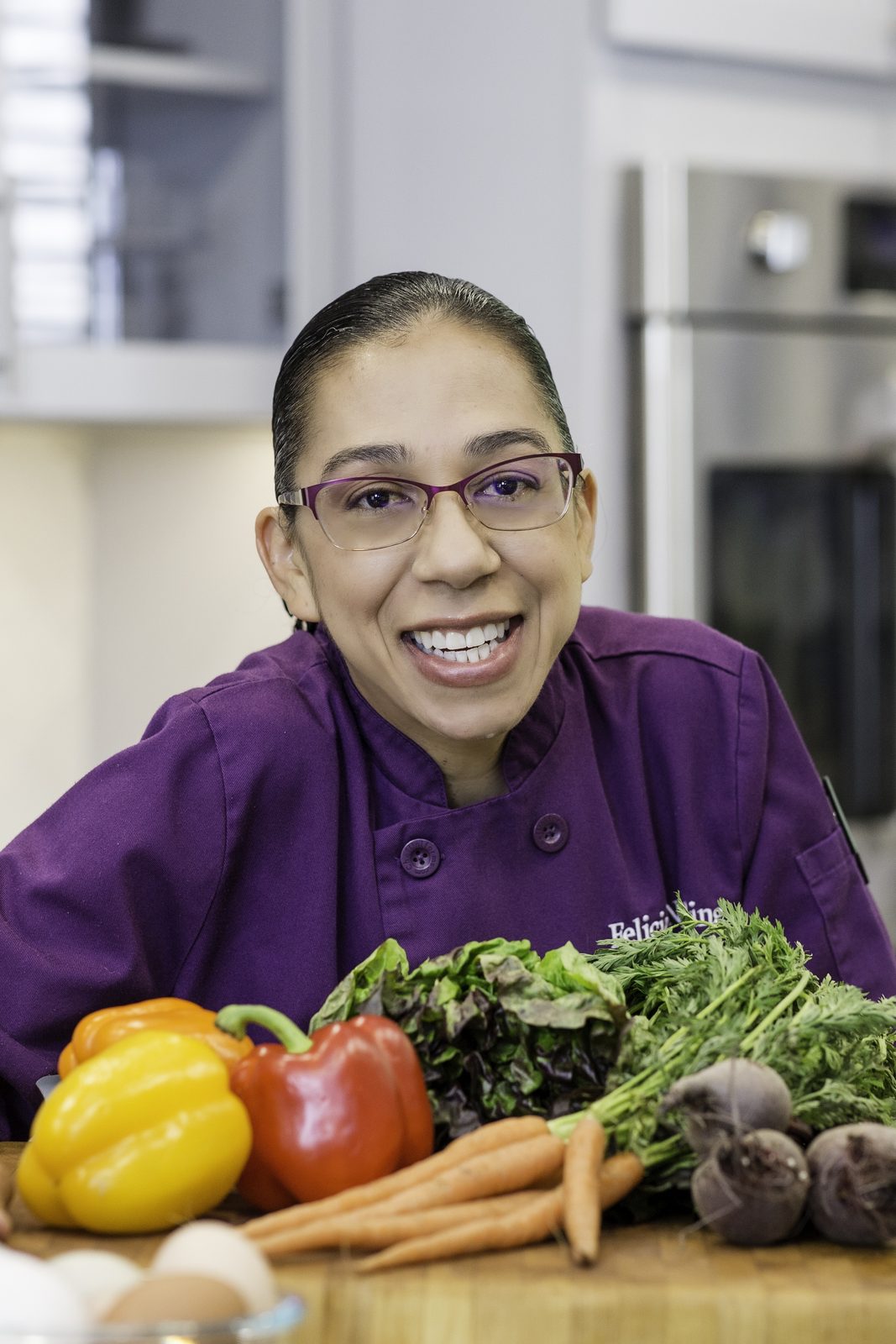 Felicia Dees, founder of Creative Meals by Chef Felicia, in a purple chef coat behind a table of fresh vegetables