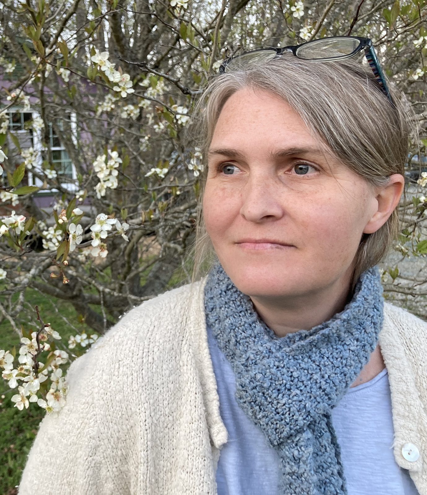 Gabrielle Lazarus, founder of Sacredsong Birth & Wellness, in a cream cardigan and knit scarf beneath a flowering tree