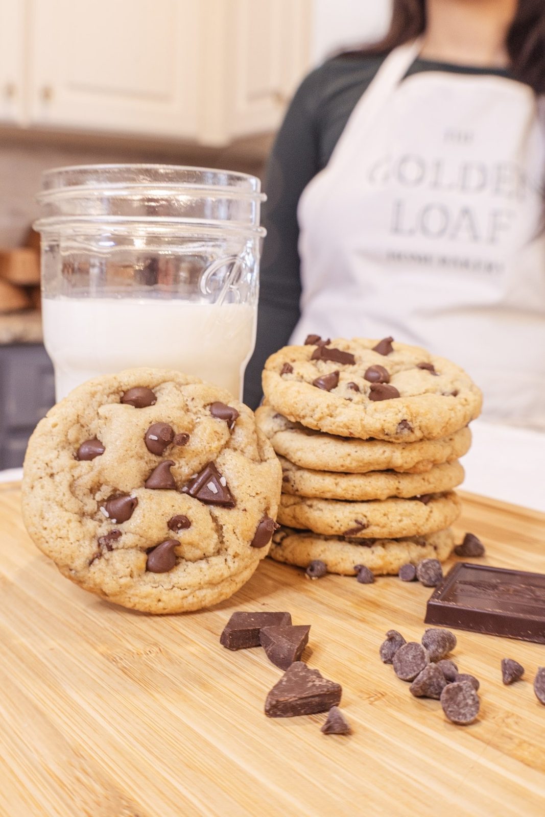 A stack of chocolate chip cookies with a jar of milk and chunks of chocolate on a wooden board