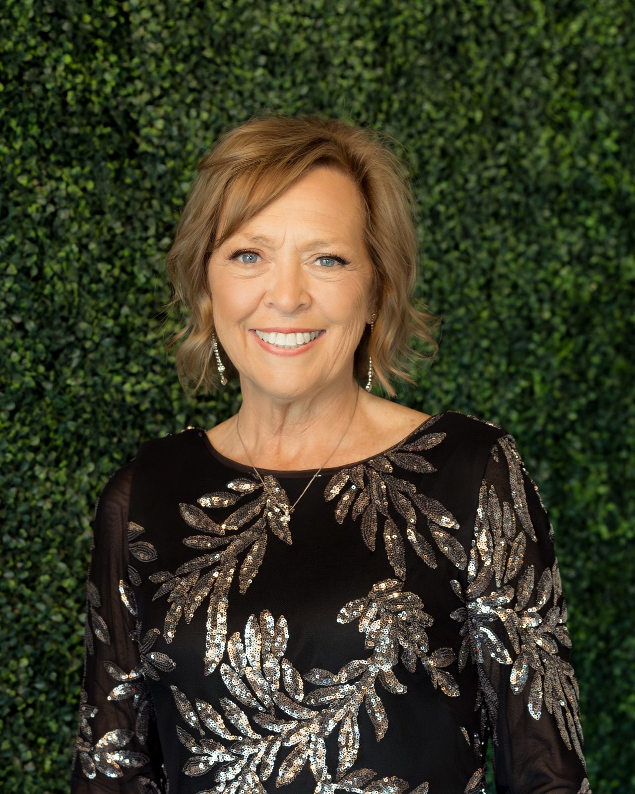 Goldie Benz, owner of GoldenWorks Embroidery, smiling in a black top with silver sequined leaves against a green hedge wall