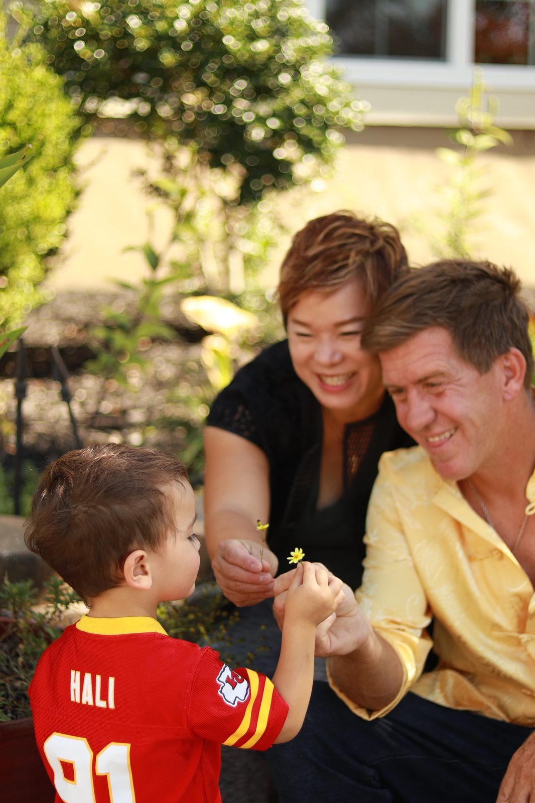 Grace Liu laughing with her husband and young son, who holds up a small yellow flower in a Kansas City Chiefs jersey