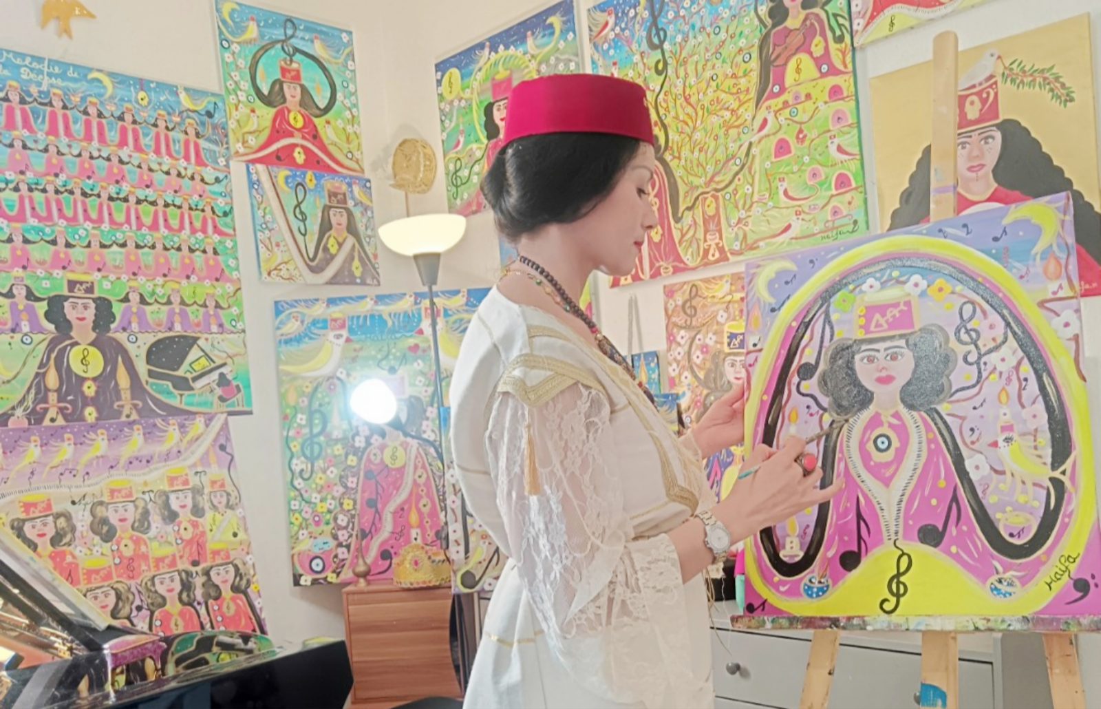 Haïfa Melliti in a white lace dress and red chechia, painting a Goddesses of Light canvas in her studio surrounded by her colourful paintings