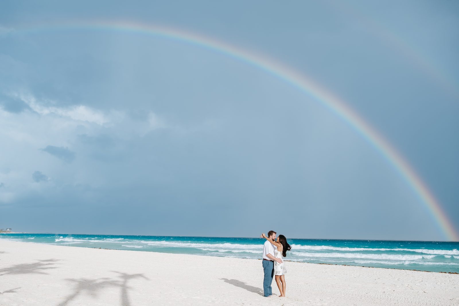 A couple embracing on a Cancún beach beneath a double rainbow, photographed by Idaly Bustamante