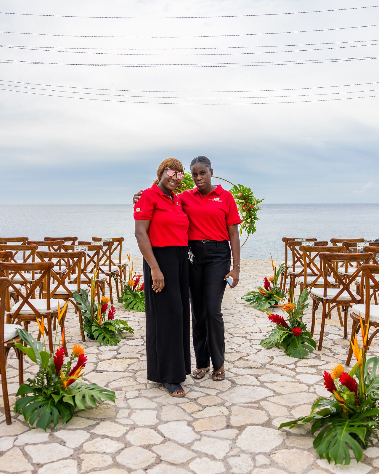 Two Weddings in Paradise planners in red team polos standing on the stone aisle of an oceanfront ceremony setup lined with tropical flowers