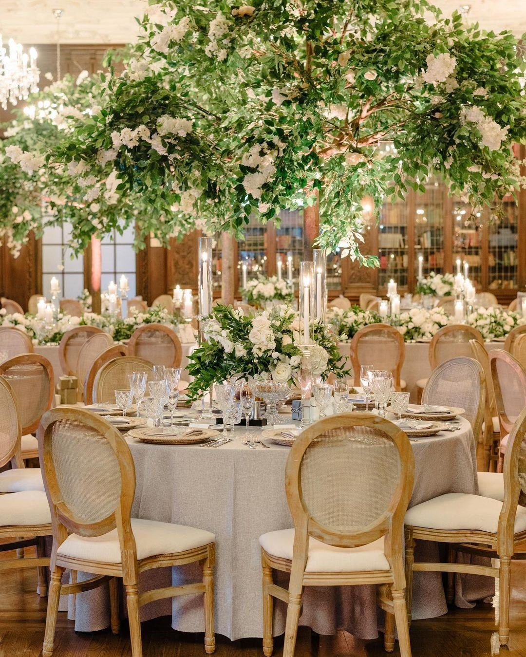 A wedding reception table set with white linens, gold-rimmed glassware and towering centerpieces of white flowers and greenery
