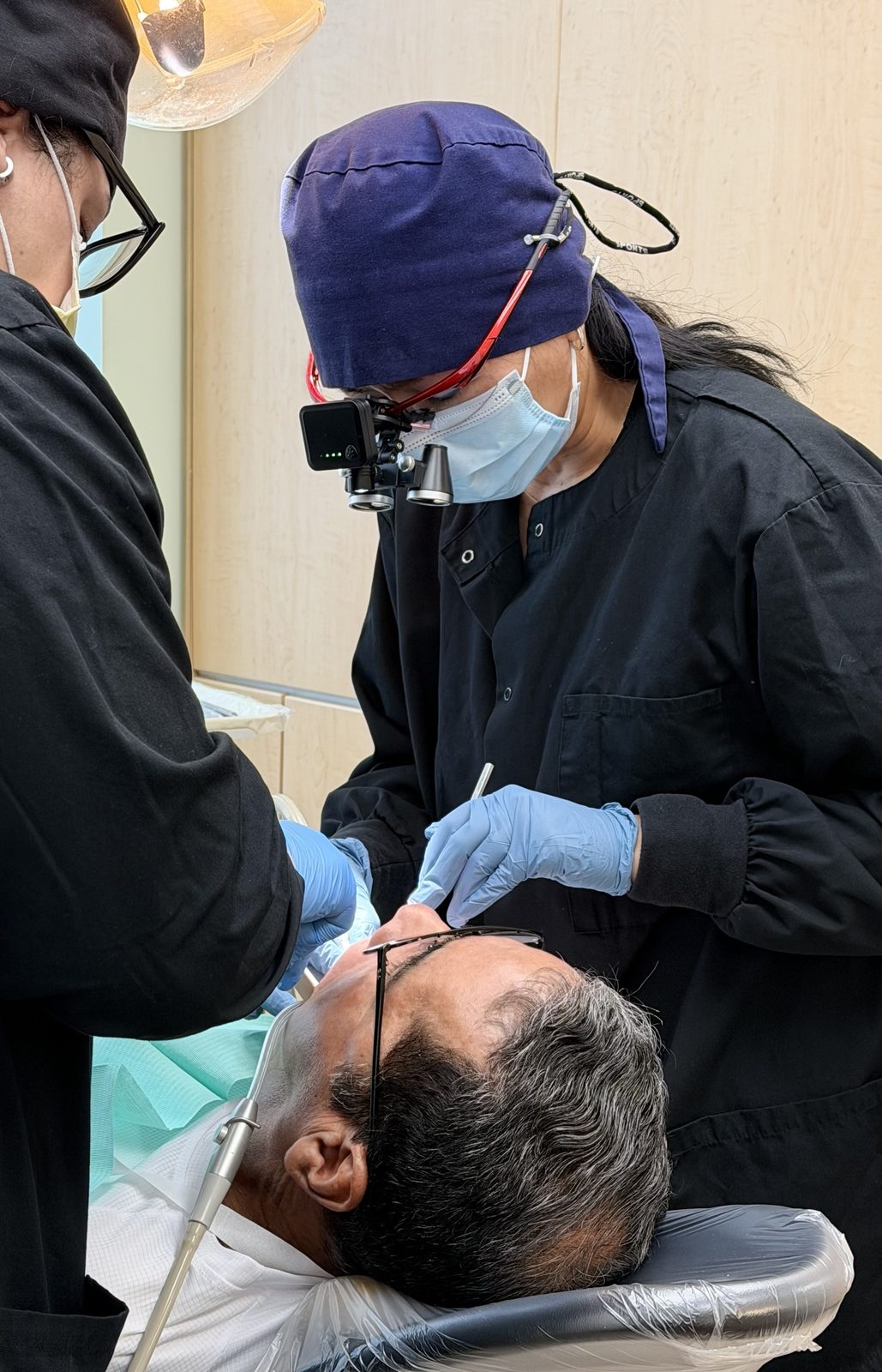 Dr. Johnimel Bianco in a navy scrub cap, mask and magnifying loupes treating a patient in the dental chair while an assistant helps