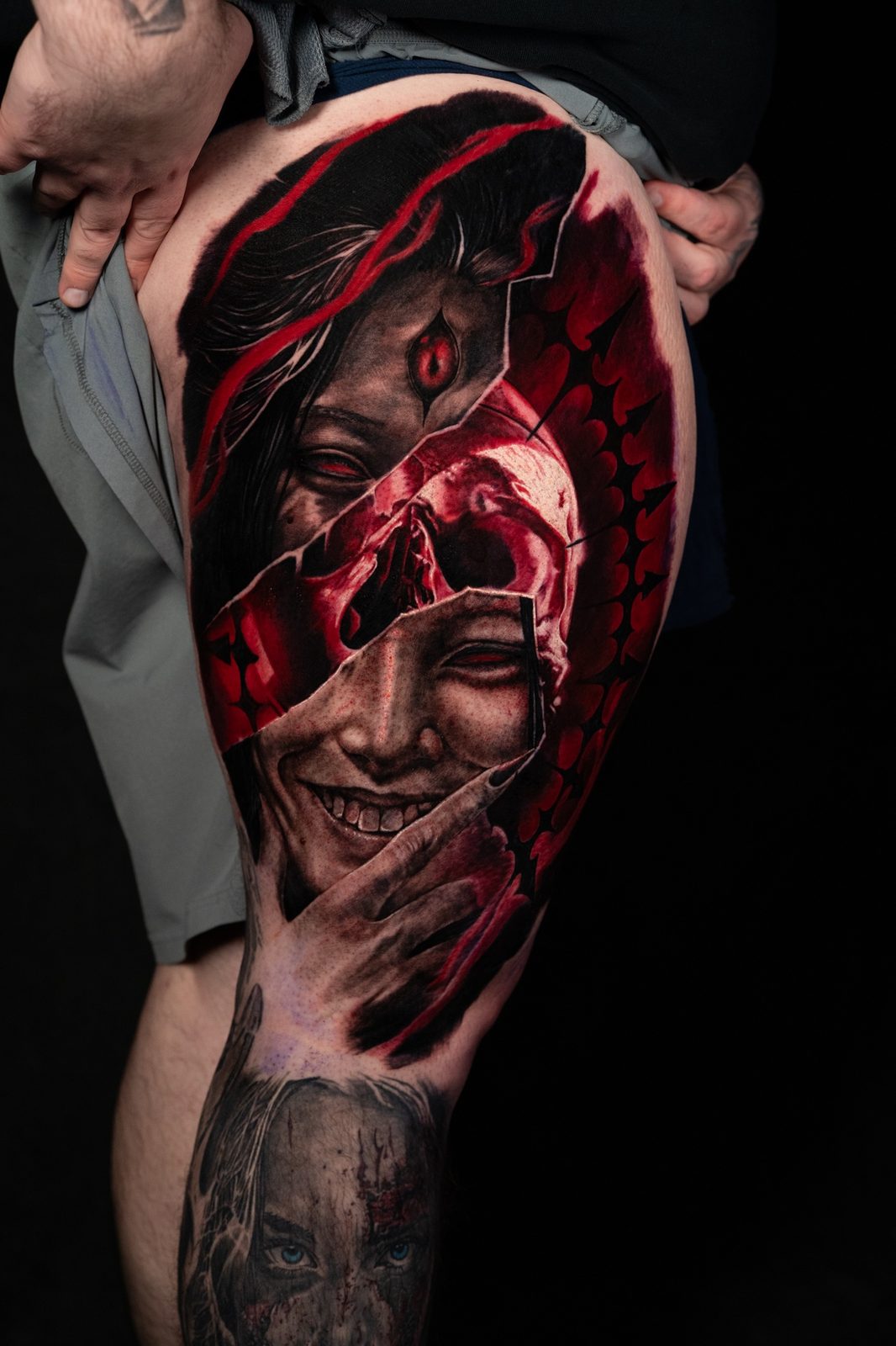 A large dark art surrealism thigh tattoo by Philly Joe in red and black, with a skull and two faces
