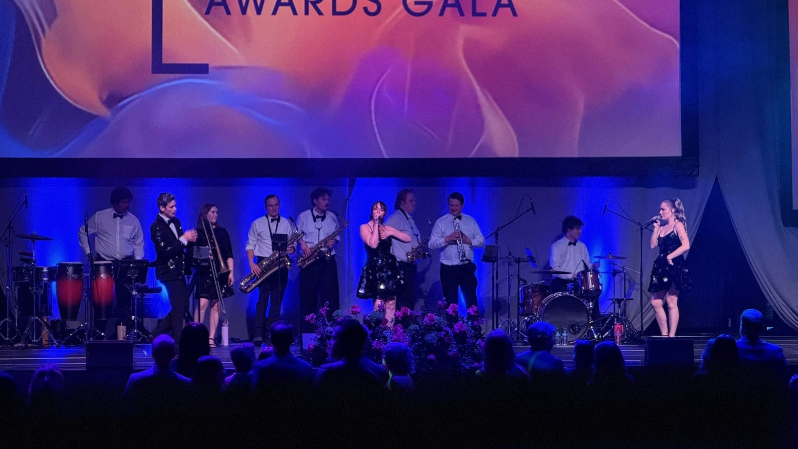 A full band with horns and vocalists performing on stage at an awards gala, Josh Luke on drums at the back