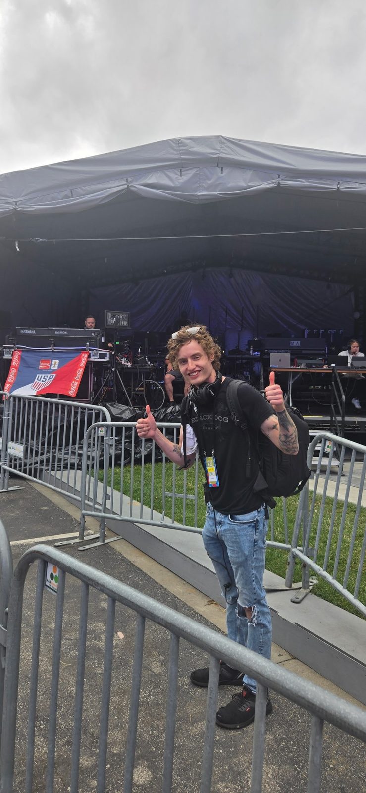 Josh Luke giving two thumbs up in front of an outdoor festival stage, wearing headphones and a backpack